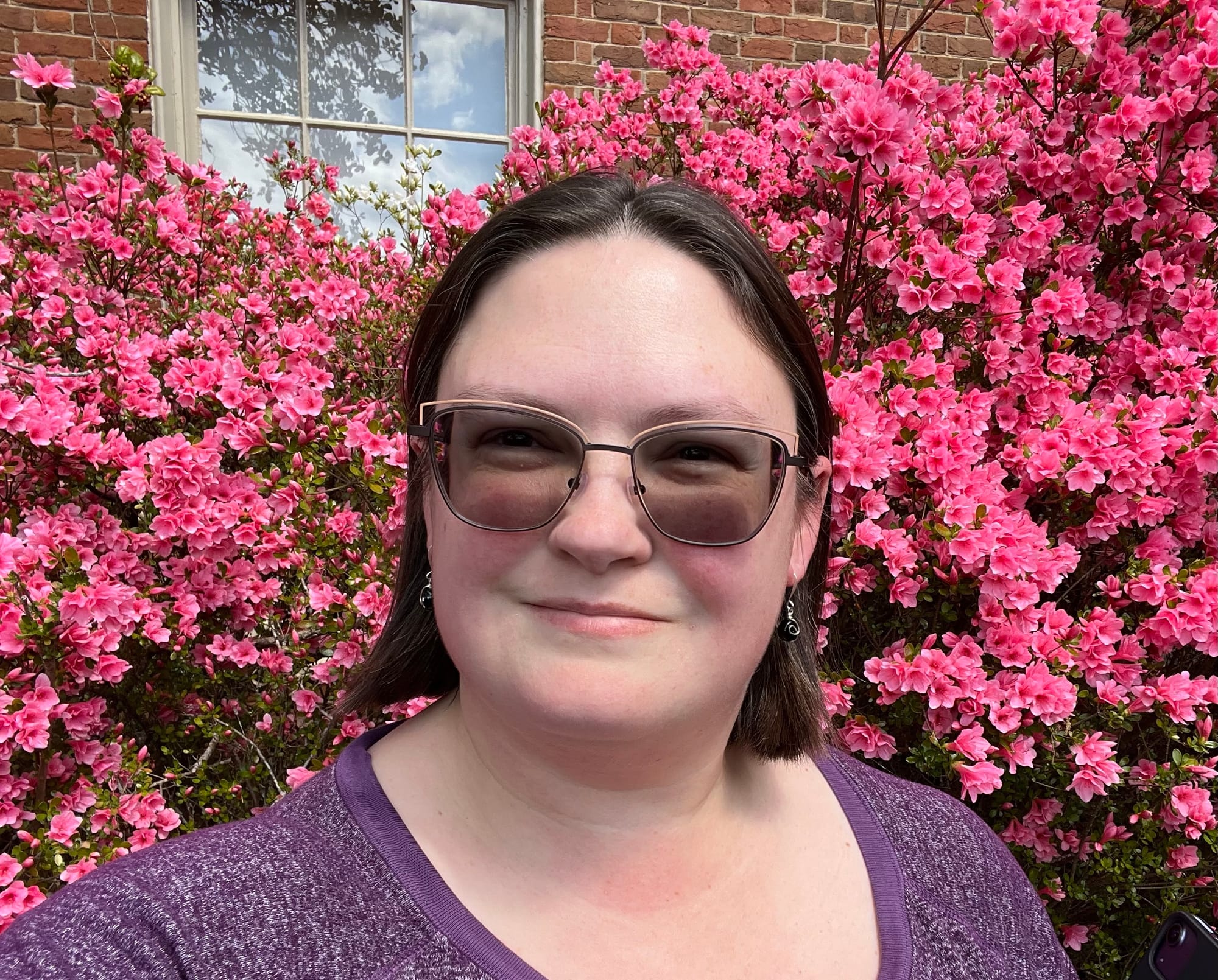 A middle-aged white woman with shoulder-length brown hair and glasses standing in front of a pink azalea bush.