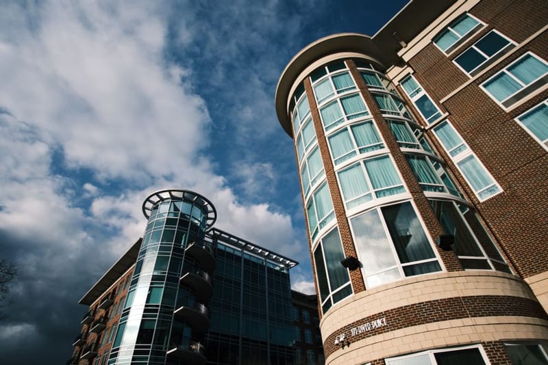 Brick buildings with bluish-green glass windows viewed from below with a blue sky full of white clouds behind them.